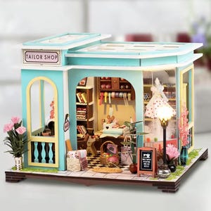 Peut inclure: Maquette de mercerie miniature avec un extérieur turquoise et blanc. Le modèle présente un intérieur détaillé avec des fournitures de couture, une machine à coudre et un sol en damier. Le panneau supérieur indique "TAILOR SHOP".