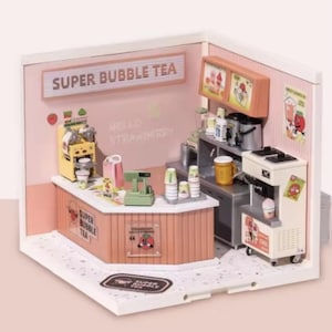 Op de afbeelding: Miniatuur diorama van een bubble tea shop, in roze en wit. De shop heeft een toonbank, drankmachines en borden met de tekst "SUPER BUBBLE TEA" en "HELLO STRAWBERRY". Er zijn kleine bekers, een kassa en andere accessoires.