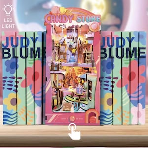 May include: A detailed miniature candy store diorama with LED lighting, featuring a rainbow and the words "CANDY STORE." The diorama is flanked by colourful book spines with the name "JUDY BLUME" and floral designs. A touch icon is visible.