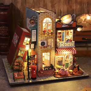 Puede incluir: Maqueta en miniatura iluminada de una escena callejera de estilo europeo. El modelo presenta una librería, un café y otras tiendas, con fachadas de ladrillo y estuco. La escena incluye una farola, un banco y un buzón rojo. La escena está iluminada con luces cálidas.