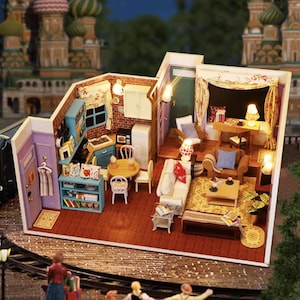 Può includere: Una casa delle bambole in miniatura dettagliata con cucina e soggiorno. La cucina è dotata di mobili blu, un fornello e un frigorifero. Il soggiorno ha un divano, un tavolino e delle lampade. La casa delle bambole è ambientata sullo sfondo di un edificio con cupole colorate.