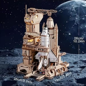 May include: A wooden space shuttle model with a launch tower, measuring 25 cm long, 19 cm wide, and 26 cm tall. The model is set against a backdrop of a moon and stars, with the dimensions labeled on the image.