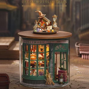 Puede incluir: Un diorama en miniatura detallado dentro de un tarro de cristal. El diorama presenta una tienda con el texto "HONEYMAGIC" sobre la ventana. Una tapa de madera sostiene un sombrero de bruja, un reloj de arena y otros objetos pequeños. El tarro se encuentra sobre una superficie adoquinada.