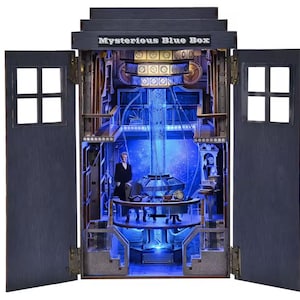 Peut inclure: Modèle détaillé d'une boîte bleue avec l'inscription "Mysterious Blue Box" en haut. L'intérieur est éclairé en bleu, révélant une figurine, une table et des mécanismes complexes. Les portes sont ouvertes, révélant l'intérieur détaillé.