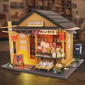 Pode incluir: Modelo em miniatura de uma loja japonesa com telhado preto e paredes bege. O modelo apresenta um letreiro em japonês, prateleiras repletas de produtos em miniatura e uma pequena caixa de correio azul. Um letreiro indica "Aberto".