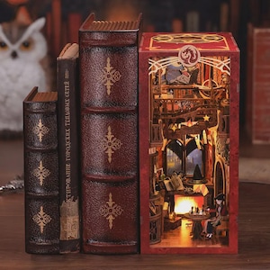 Puede incluir: Una estantería decorativa con tres libros encuadernados en símil piel de diferentes tamaños. Los libros son de color rojo oscuro con diseños dorados en relieve. Un diorama detallado e iluminado de una habitación es visible dentro del libro de la derecha, con una chimenea y figuras.