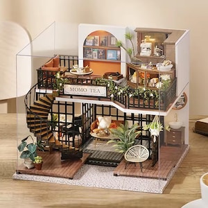 Op de afbeelding: Een miniatuur poppenhuiscafé met meerdere niveaus, met een wenteltrap, balkons en gedetailleerde interieurdecoratie. Het café heeft de tekst "MOMO TEA" op een bord. De structuur omvat miniatuurmeubels, planten en decoratieve items.