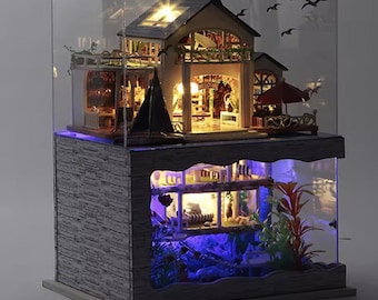 May include: A miniature dollhouse with interior lighting, enclosed in a clear case. The two-story structure features a balcony, a small sailboat, and a red umbrella. The base has a faux stone design with blue LED lighting, creating a cozy, illuminated scene.