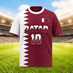 K&ouml;nnte beinhalten: Bordeaux-wei&szlig;es Fu&szlig;balltrikot mit dem Wort "QATAR" und der Nummer "10" auf der Vorderseite. Das Trikot hat ein wei&szlig;es Zickzack-Design auf der linken Seite und eine kleine Katar-Flagge auf der rechten Brust. Das Trikot hat einen wei&szlig;en Kragen und &Auml;rmelbesatz.