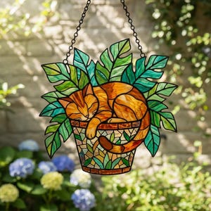 Könnte beinhalten: Buntglas-Fensterdekoration mit einer schlafenden, orangefarbenen Katze in einem Mosaik-Blumentopf, umgeben von grünen Blättern. Die Katze ist zusammengerollt, und der Topf wird von einer schwarzen Kette gehalten. Ein dekoratives Stück für Fenster oder Garten.