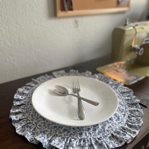 Puede incluir: Un plato blanco con un tenedor y una cuchara plateados descansa sobre un mantel individual floral azul con un borde con volantes. El mantel individual y el plato están sobre una superficie de madera marrón oscuro. El mantel individual tiene un delicado estampado floral azul.