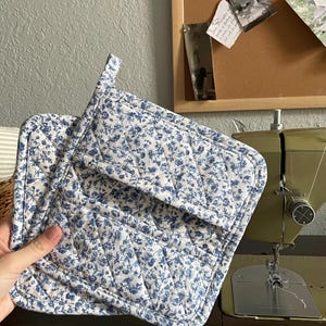 May include: A pair of quilted pot holders with a white background and a blue floral pattern. The pot holders are square with a loop for hanging. The stitching is visible, and the edges are bound with a matching fabric.