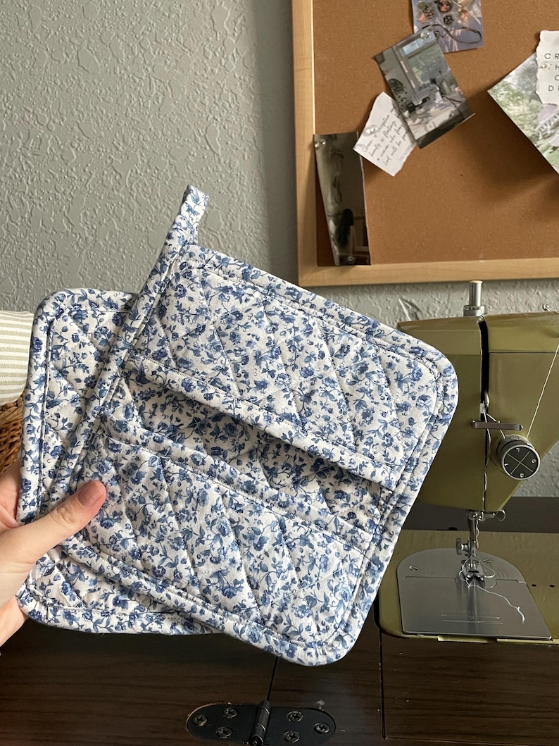 May include: A pair of quilted pot holders with a white background and a blue floral pattern. The pot holders are square with a loop for hanging. The stitching is visible, and the edges are bound with a matching fabric.