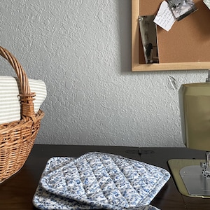 May include: A quilted pot holder with a blue floral pattern on a white background. The pot holder is folded on a dark wooden surface. A wicker basket with a striped fabric lining is in the background.