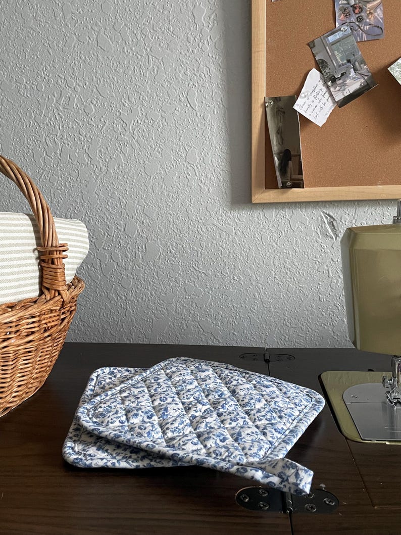 May include: A quilted pot holder with a blue floral pattern on a white background. The pot holder is folded on a dark wooden surface. A wicker basket with a striped fabric lining is in the background.