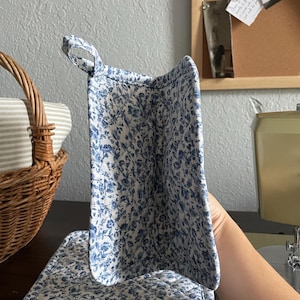 May include: A quilted pot holder and stand set with a white background and blue floral pattern. The pot holder has a loop for hanging. A wicker basket with striped fabric is in the background.