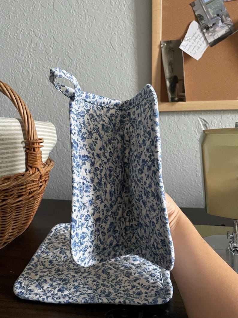 May include: A quilted pot holder and stand set with a white background and blue floral pattern. The pot holder has a loop for hanging. A wicker basket with striped fabric is in the background.