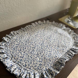 May include: An oval placemat featuring a blue floral pattern on a white background. It has a ruffled edge for a decorative finish. The fabric is likely cotton or a similar material. The placemat is designed for table settings.