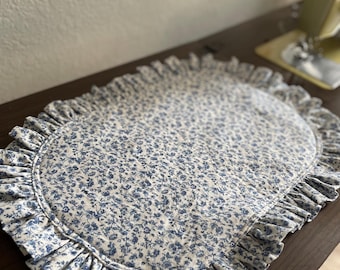 May include: An oval-shaped placemat with a white background and a blue floral pattern. The placemat has a ruffled edge, adding a decorative touch. The fabric appears to be cotton or a similar material.