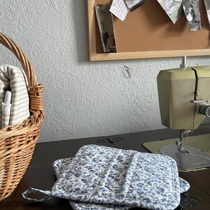 May include: Two blue floral quilted pot holders with a loop for hanging rest on a dark wooden surface. A vintage green sewing machine is visible in the background, along with a wicker basket and a corkboard with photos.