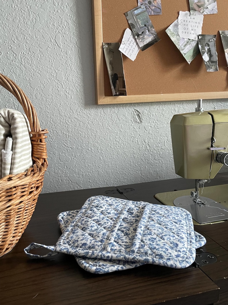 May include: Two blue floral quilted pot holders with a loop for hanging rest on a dark wooden surface. A vintage green sewing machine is visible in the background, along with a wicker basket and a corkboard with photos.