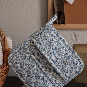 Puede incluir: Un juego de agarradera y salvamanteles acolchados. La tela presenta un fondo blanco con un delicado estampado floral azul. La agarradera tiene un lazo para colgar. El juego está diseñado para uso en la cocina.