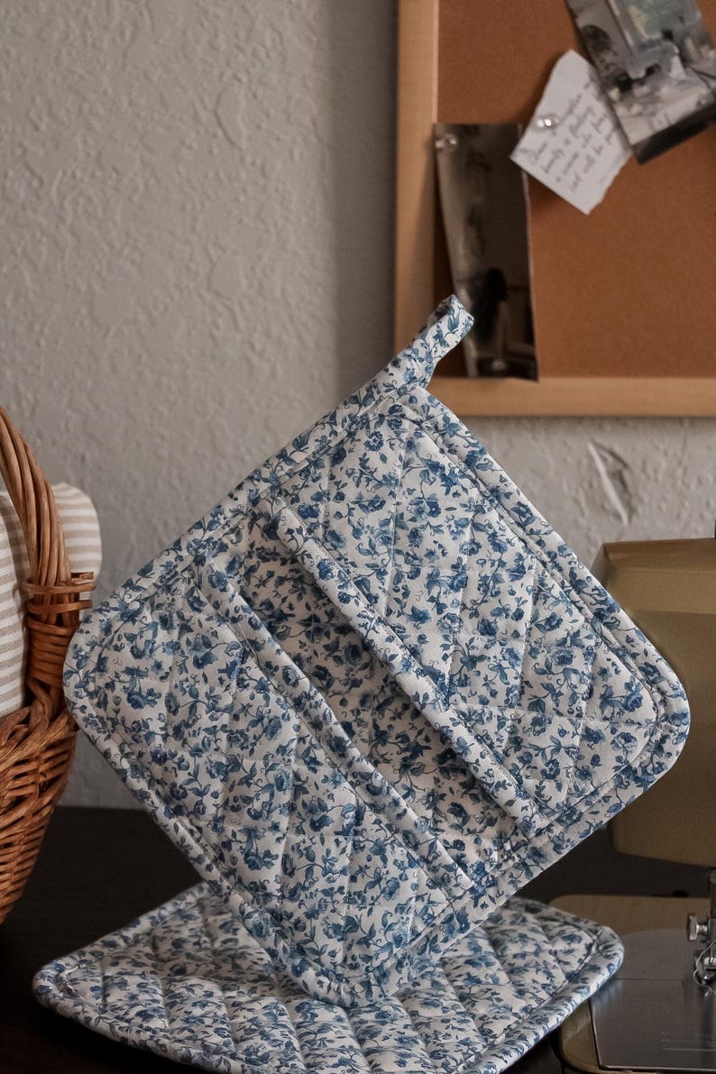 May include: A quilted pot holder and matching trivet set. The fabric features a white background with a delicate blue floral pattern. The pot holder has a loop for hanging. The set is designed for kitchen use.