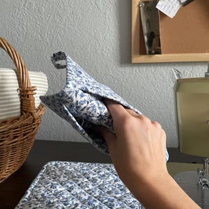 May include: A quilted oven mitt and matching hot pad set. Both items feature a blue floral pattern on a white background. The mitt is held up, showing the interior. The hot pad is on a dark surface.