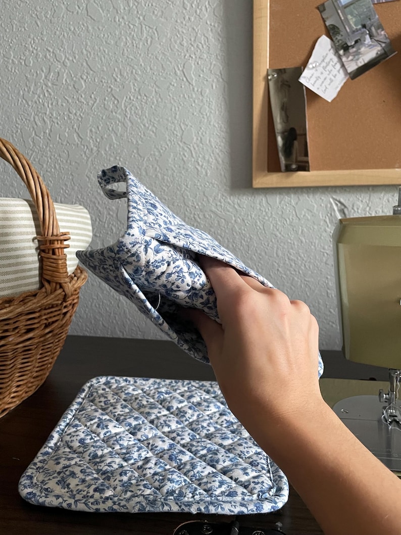 May include: A quilted oven mitt and matching hot pad set. Both items feature a blue floral pattern on a white background. The mitt is held up, showing the interior. The hot pad is on a dark surface.