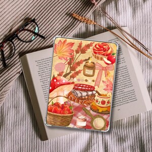 May include: A rectangular tablet case featuring a warm-toned autumn illustration with a basket of red apples, a large mushroom, a candle, a plaid blanket, a tart, a red bow, and a hand mirror, all set against a textured, light-coloured background.