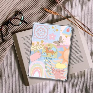 Può includere: Una custodia trasparente per tablet con un design colorato e stravagante con un sole, stelle, fiori, una farfalla, un ariete e un axolotl. La parola "ARIES" è visualizzata. La custodia è appoggiata su un libro aperto.