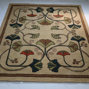 May include: A rectangular area rug with a beige background featuring a symmetrical floral pattern. The design includes stylized leaves and blossoms in shades of dark blue, olive green, and burnt orange, enclosed by a dark brown border.