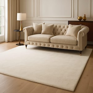 May include: A cream-colored rug and sofa set in a living room setting. The tufted sofa has three matching pillows and wooden legs. A gold lamp and side table are to the left of the sofa. The room has hardwood floors and a neutral color palette.