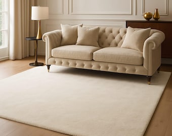 May include: A cream-colored rug and sofa set in a living room setting. The tufted sofa has three matching pillows and wooden legs. A gold lamp and side table are to the left of the sofa. The room has hardwood floors and a neutral color palette.