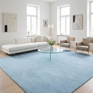 Könnte beinhalten: Ein modernes Wohnzimmer mit einem hellblauen Teppich, einem weißen Sofa und zwei beigefarbenen Sesseln. Ein Glastisch und abstrakte Kunstwerke ergänzen das minimalistische Design. Der Raum hat große Fenster und Holzböden.