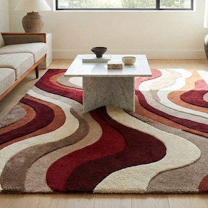 Könnte beinhalten: Ein großer Teppich mit einem wellenförmigen Muster in Rot-, Braun-, Beige- und Orangetönen. Der Teppich liegt auf einem Holzboden in einem Wohnzimmer, mit Sofa und Couchtisch. Maße: ca. 200 cm x 300 cm.