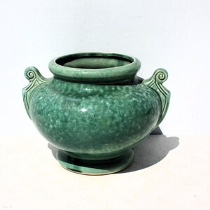 May include: A green ceramic vase with a speckled texture. The vase has a rounded body, a wide opening, and two decorative handles. It sits on a small, circular base. The overall design is vintage and decorative.