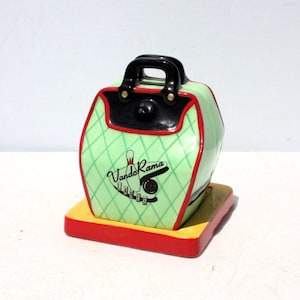 May include: A ceramic bowling bag-shaped cookie jar featuring a light green body with a dark green diamond grid pattern, a black handle, and red trim. The front displays a black graphic with a bowling pin and ball, reading VandoRama LANES.