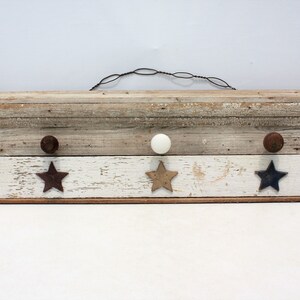 May include: Rustic wooden wall shelf with three hooks, featuring a white knob, two brown knobs, and three star-shaped accents in brown, gold, and blue.