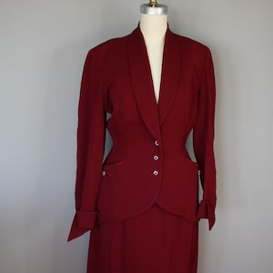 May include: A burgundy vintage suit with a skirt and a jacket. The jacket has a shawl collar and a single button closure. The skirt is knee-length and has a straight silhouette.