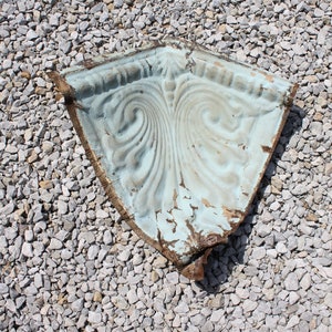 May include: A weathered, light blue metal corner piece with a decorative, swirling design. The piece is lying on a bed of small, gray gravel.