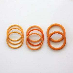 May include: Three sets of orange plastic bangles. The bangles are stacked on top of each other, with the largest bangle on the bottom and the smallest bangle on top. The bangles are all different shades of orange.