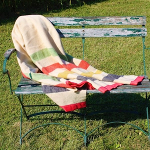 May include: A striped blanket in red, yellow, green, and white is draped over a green metal bench with a wooden seat. The bench is outdoors on a grassy lawn.