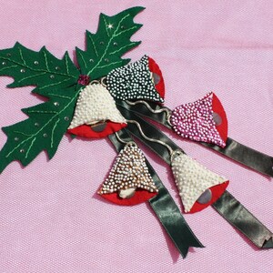 May include: A green felt holly leaf with four beaded bells attached. The bells are white, pink, gold, and green with red felt tops. The bells are attached to black satin ribbons.