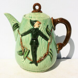 May include: Green ceramic teapot with a brown handle and a whimsical design of a figure in green clothing climbing a vine. The teapot has a green and white striped pattern.