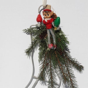 May include: A metal stocking holder with a green and red Christmas lights and a small elf figurine sitting inside. The stocking holder is decorated with evergreen branches.