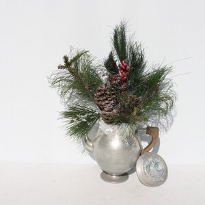 May include: A silver teapot with a wooden handle, filled with pine branches, pine cones, and red berries. The teapot is sitting on a white surface.