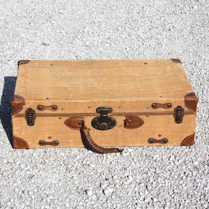 May include: A rectangular tan suitcase with a textured exterior, featuring brown leather corner reinforcements, a central metal latch with a keyhole, two side metal buckles, and a brown leather handle attached to the front panel.