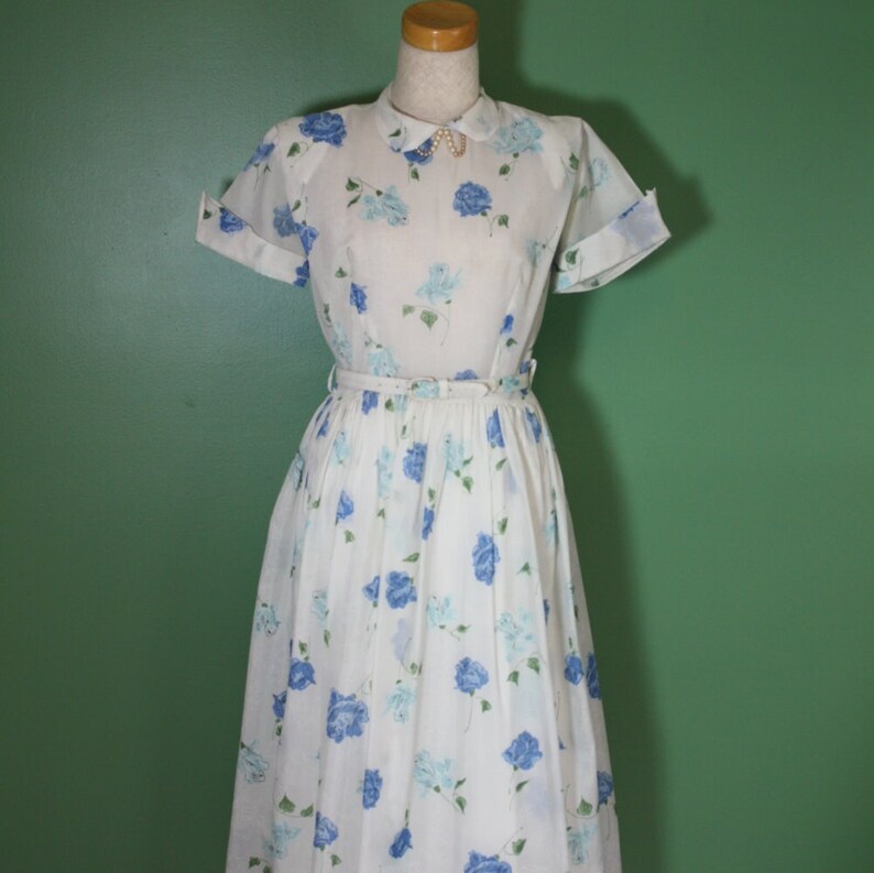 May include: A white dress with a blue floral print and a white collar. The dress has short sleeves and a fitted waist. The dress is on a mannequin against a green background.