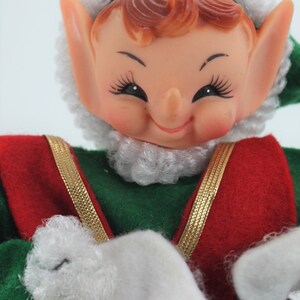 May include: A smiling elf doll with a white fur hat and beard, wearing a green and red outfit with gold trim. The elf is holding its hands in front of it, with white fur mittens.